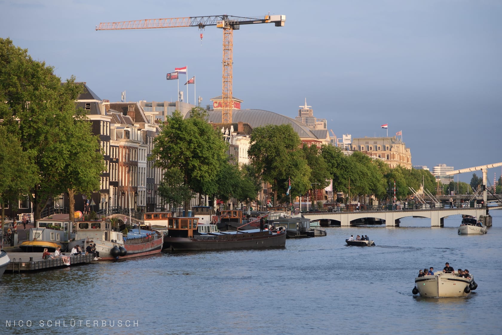 Abendlicht an der Amstel in Amsterdam mit vertäuten Hausbooten und Frachtkähnen am Kai, Ausflugsbooten auf dem Wasser und einem Baukran über der Häuserzeile