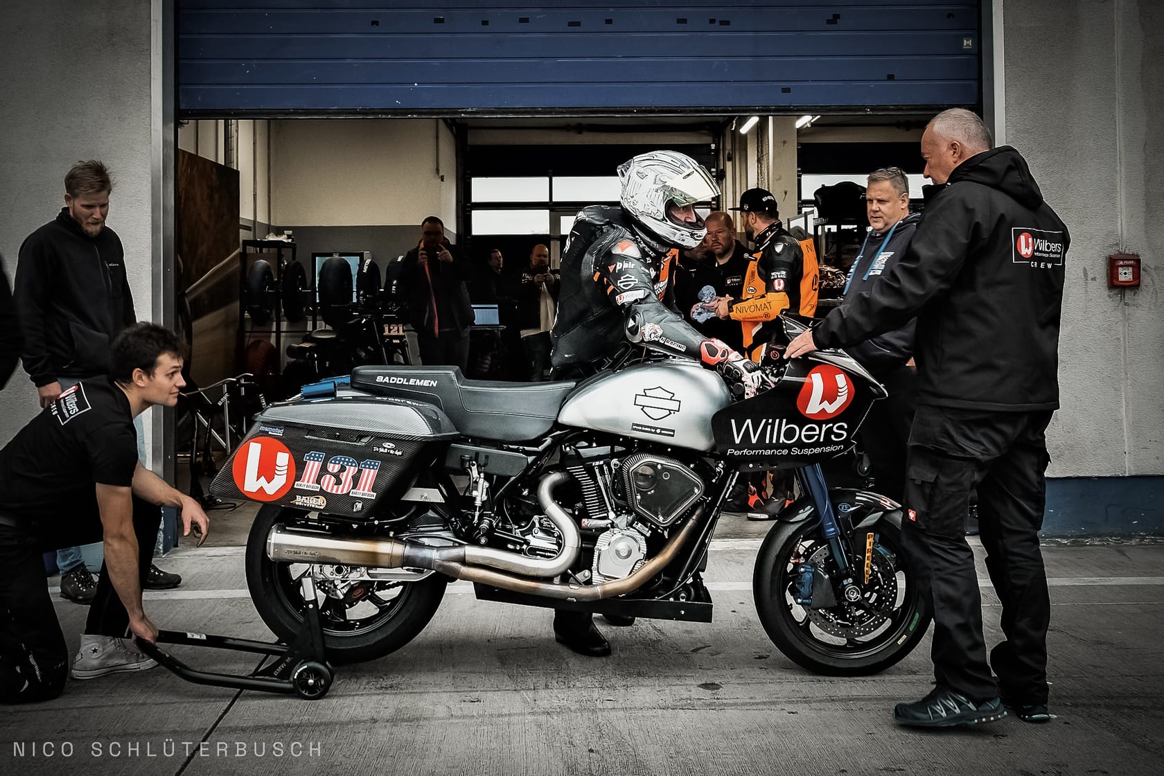 Rennfahrer mit weissem Helm steigt vor der Boxengarage auf einen silbernen Harley-Davidson Bagger-Racer mit Startnummer 131 und Wilbers-Verkleidung, umringt vom Team