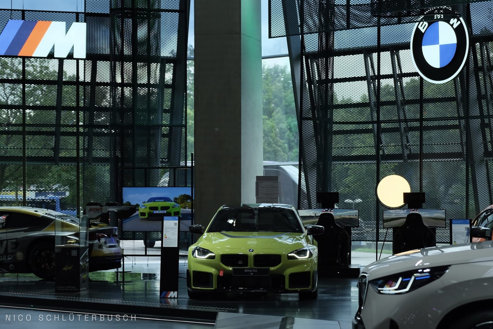 Gelbgrüner BMW M2 in der Frontansicht in einem Ausstellungsraum mit BMW- und M-Logo, umgeben von Rennsimulatoren und weiteren Fahrzeugen