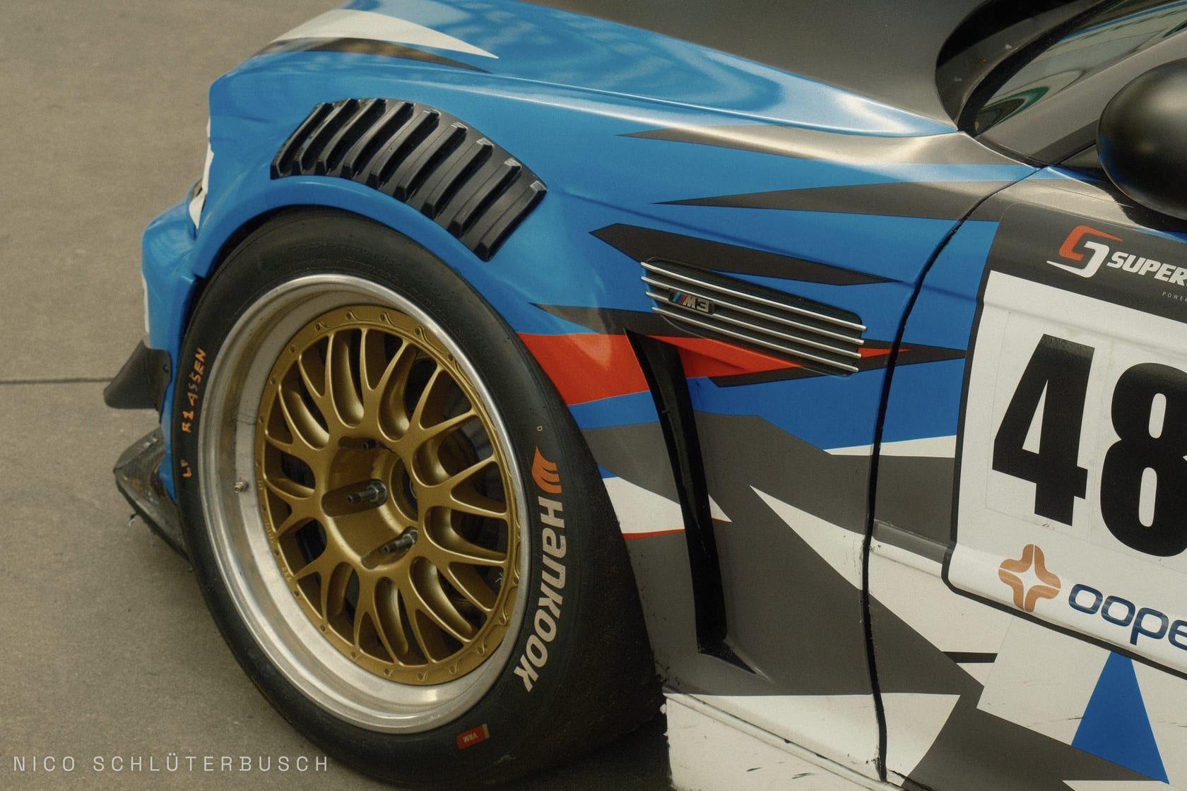 Nahaufnahme der Frontpartie eines BMW M3 Rennwagens mit Startnummer 48, blau-schwarzer Camouflage-Folierung, goldener Kreuzspeichenfelge und Hankook-Slick