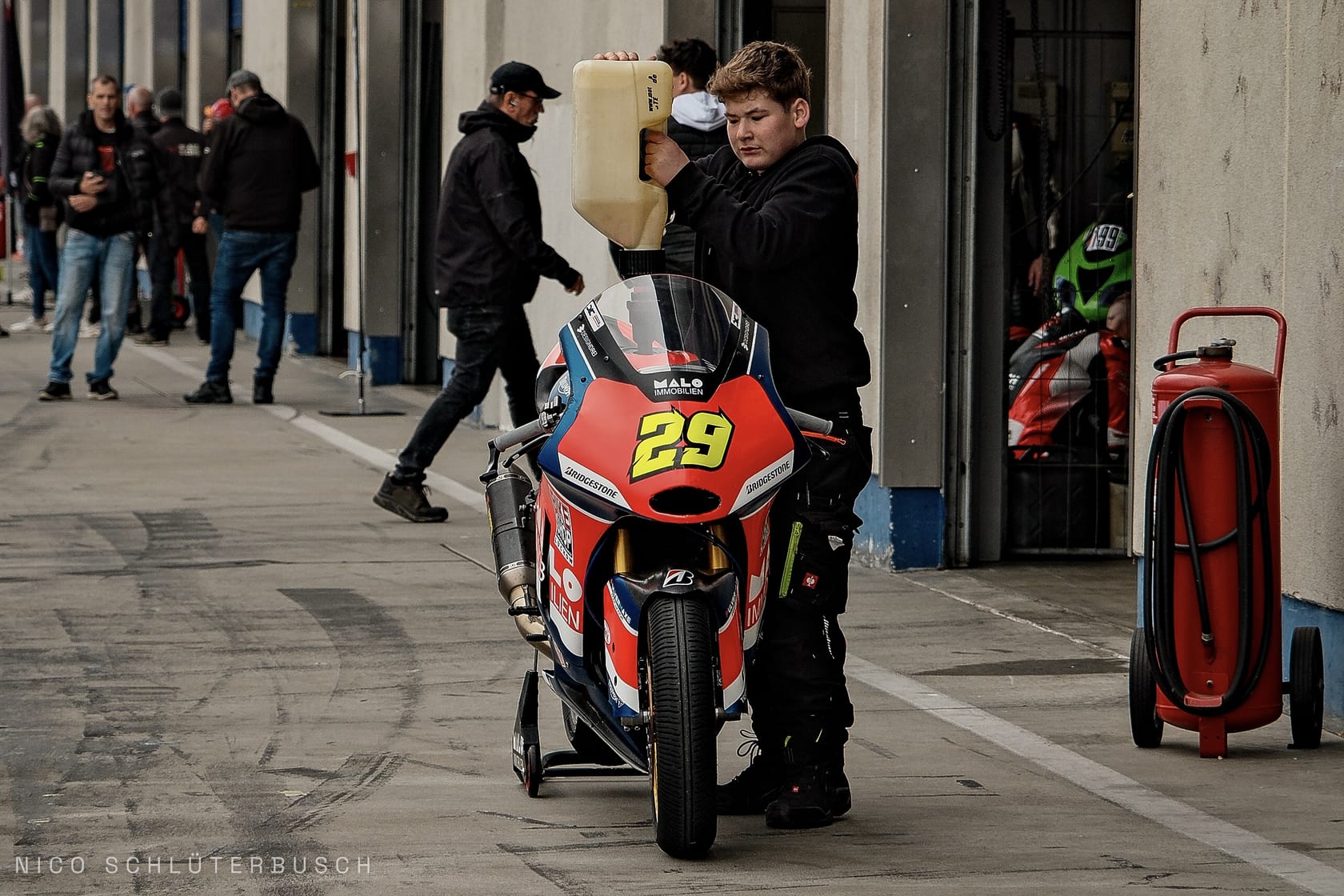 Junger Mechaniker betankt ein rot-blaues Rennmotorrad mit der Startnummer 29 aus einem Kanister in der Boxengasse