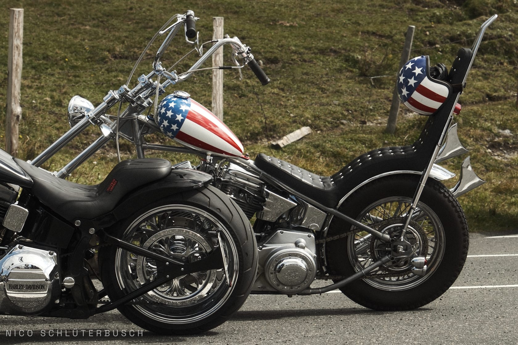 Harley-Davidson Chopper in der Seitenansicht mit Stars-and-Stripes-Tank, hoher Sissybar und passend lackiertem Helm vor einer Wiesenböschung