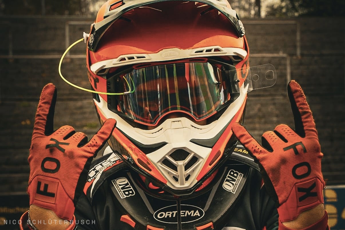 Motocross-Fahrer mit rot-weißem Helm, verspiegelter Brille und Nackenschutz zeigt mit beiden Fox-Handschuhen die Rock-Geste in die Kamera