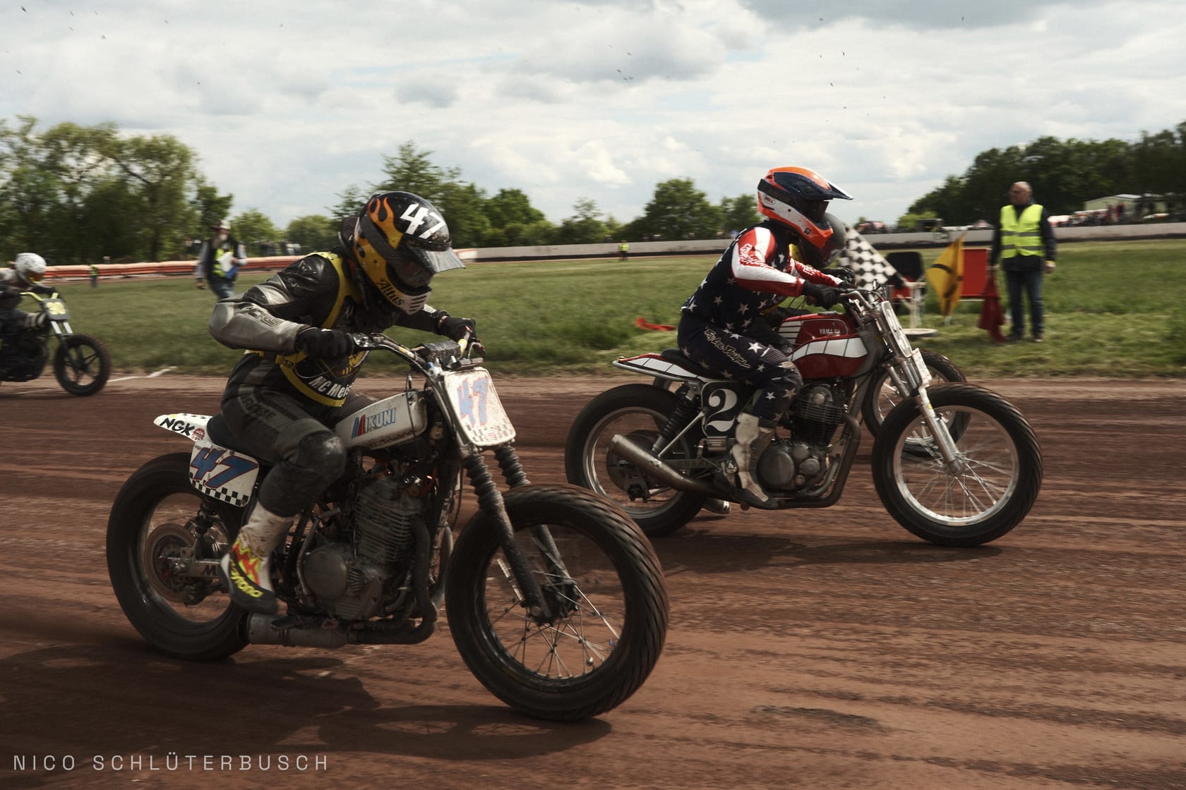Zwei Flat-Track-Fahrer im Zweikampf auf einer Dirt-Track-Bahn, vorn die Startnummer 47, dahinter ein Fahrer im Stars-and-Stripes-Dress vor der Zielflagge