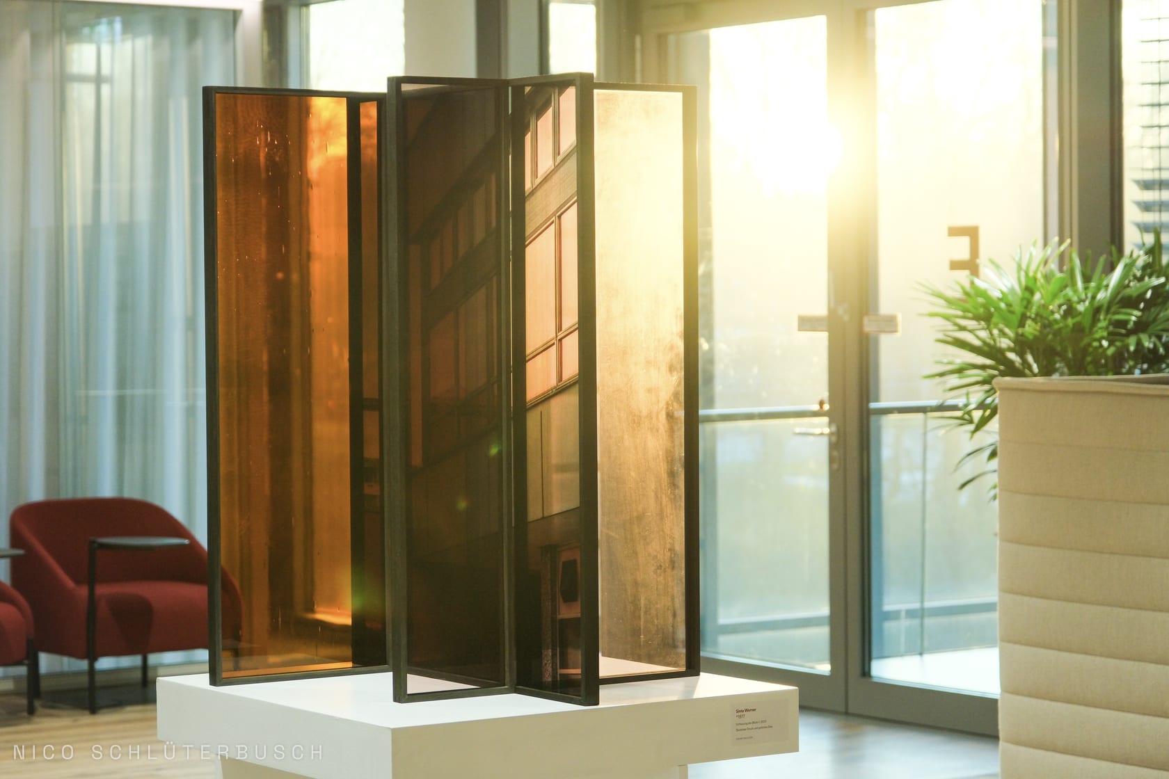 Skulptur aus getönten Glaspaneelen auf einem weißen Sockel in einem sonnendurchfluteten Foyer im Gegenlicht