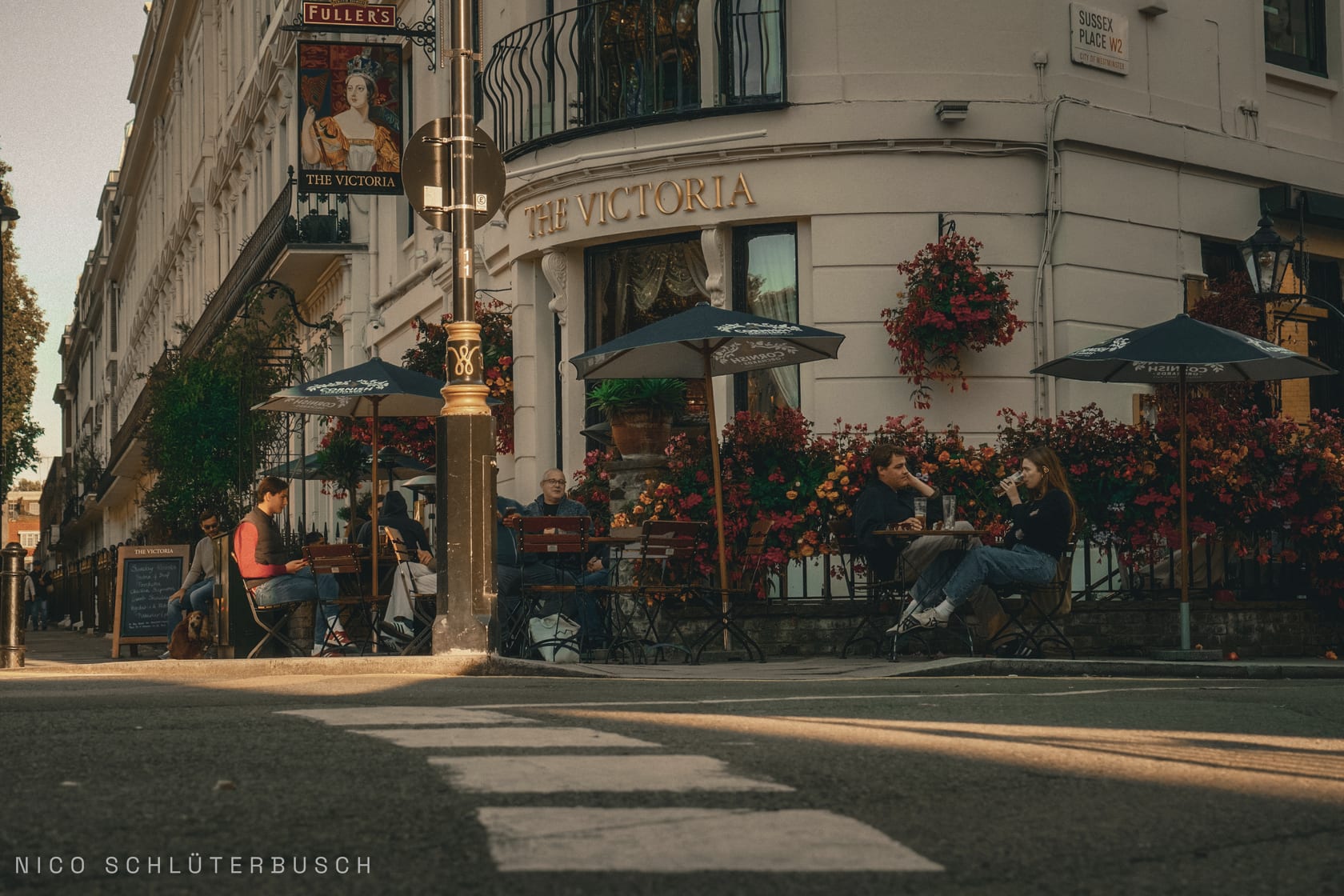 Straßenecke am Londoner Pub The Victoria mit blumengeschmückter Fassade und Gästen unter Sonnenschirmen in tiefstehender Sonne.