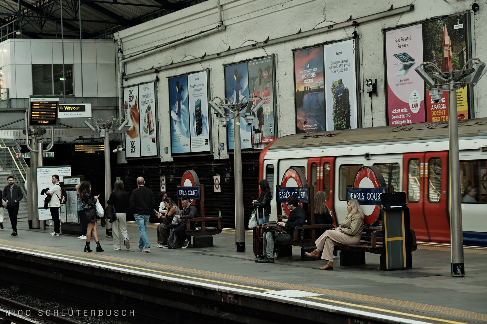 Wartende Fahrgäste auf dem Bahnsteig der Londoner U-Bahn-Station Earl's Court, rechts ein rot-weißer District-Line-Zug vor plakatierter Wand