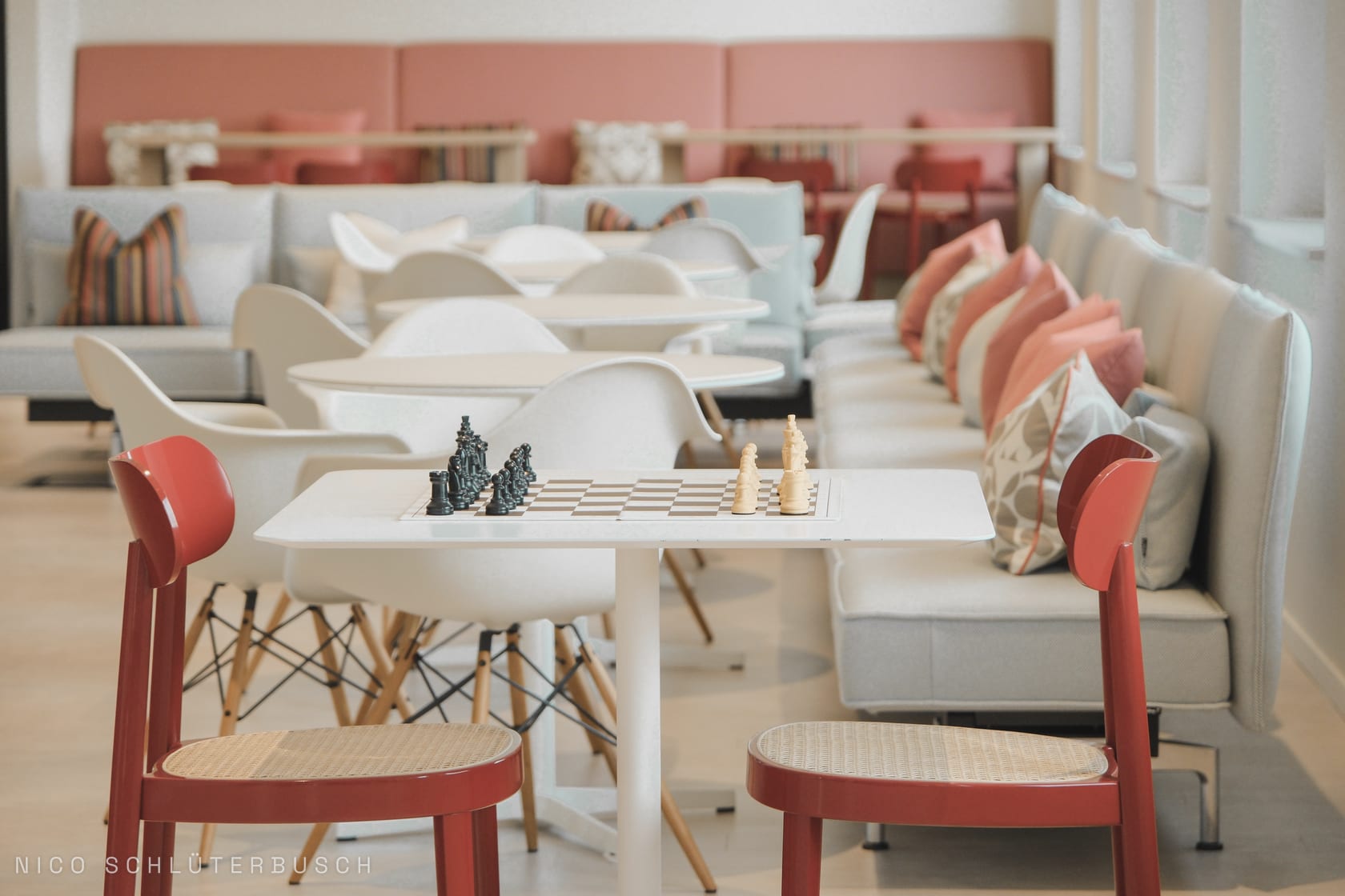 Aufgebautes Schachspiel auf einem weißen Tisch zwischen zwei roten Stühlen in einer hellen Lounge mit pastellfarbenen Polstermöbeln
