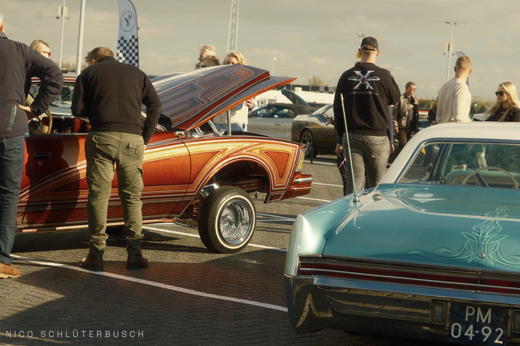 Orange-metallic lackierter Lowrider mit geöffneter Motorhaube und hochgestellter Vorderachse neben einem türkisfarbenen Oldtimer auf einem Autotreffen