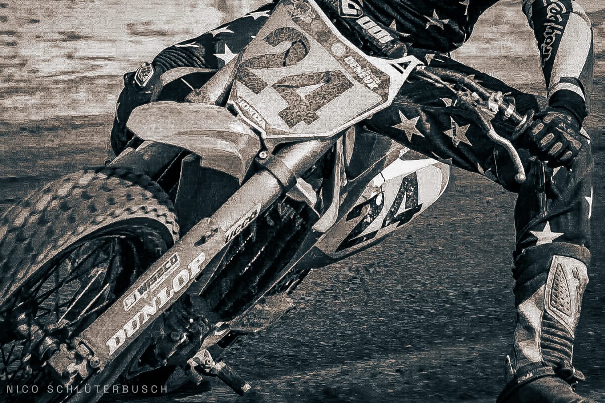 Motocross-Fahrer mit Startnummer 24 legt seine Honda in Schräglage, Nahaufnahme von Vorderrad, Gabel und ausgestrecktem Bein in Sepiatönen