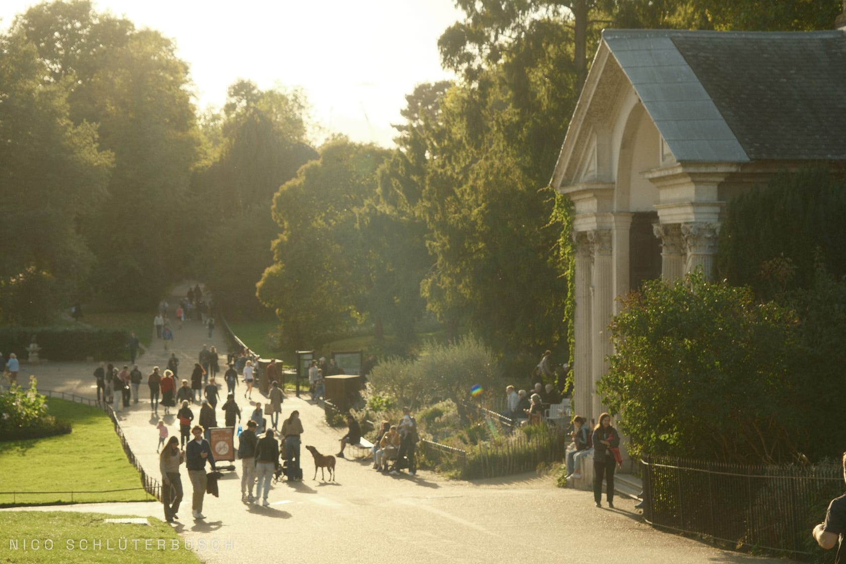 Spaziergänger auf einem breiten Parkweg im goldenen Abendlicht, rechts ein klassizistischer Tempelbau mit Säulenportikus zwischen Bäumen