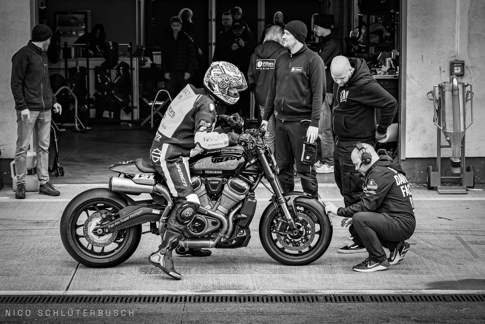 Rennfahrer in Lederkombi sitzt in der Boxengasse auf einer Harley-Davidson mit Slicks, während ein Mechaniker mit Headset kniend den Vorderreifen prüft