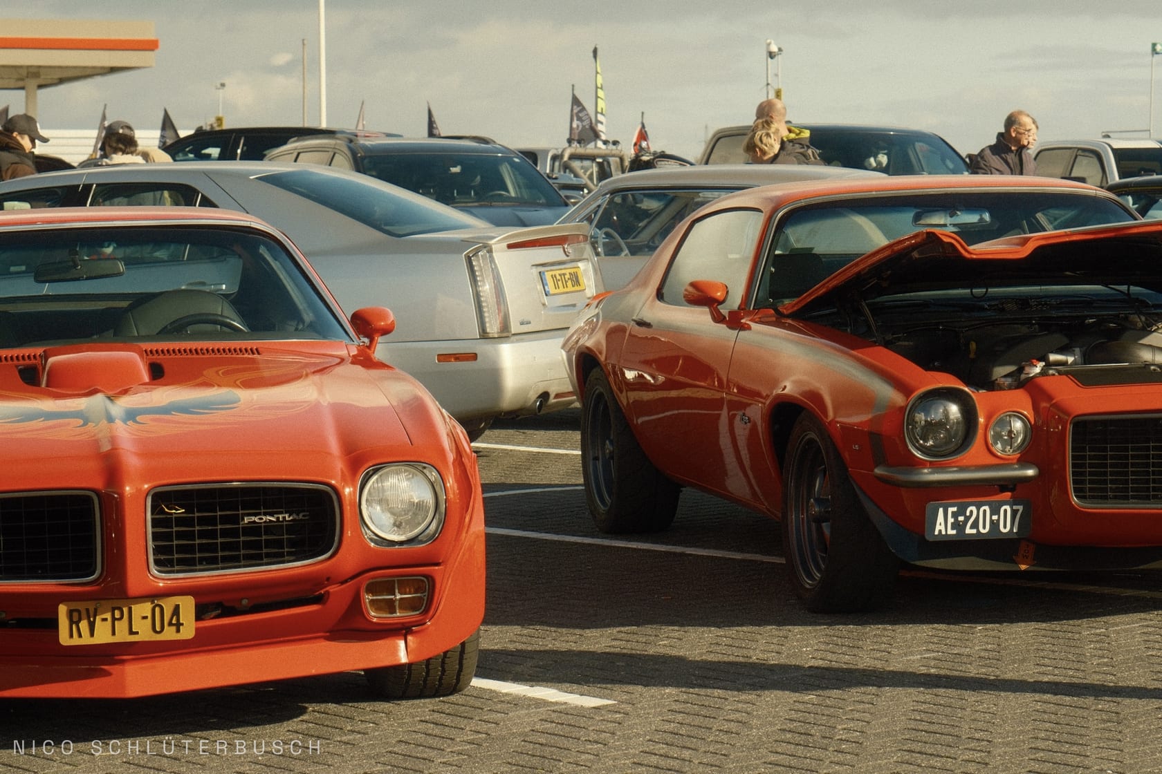 Orangeroter Pontiac Firebird und Chevrolet Camaro mit geöffneter Motorhaube nebeneinander auf einem gepflasterten Parkplatz bei einem US-Car-Treffen im Abendlicht.
