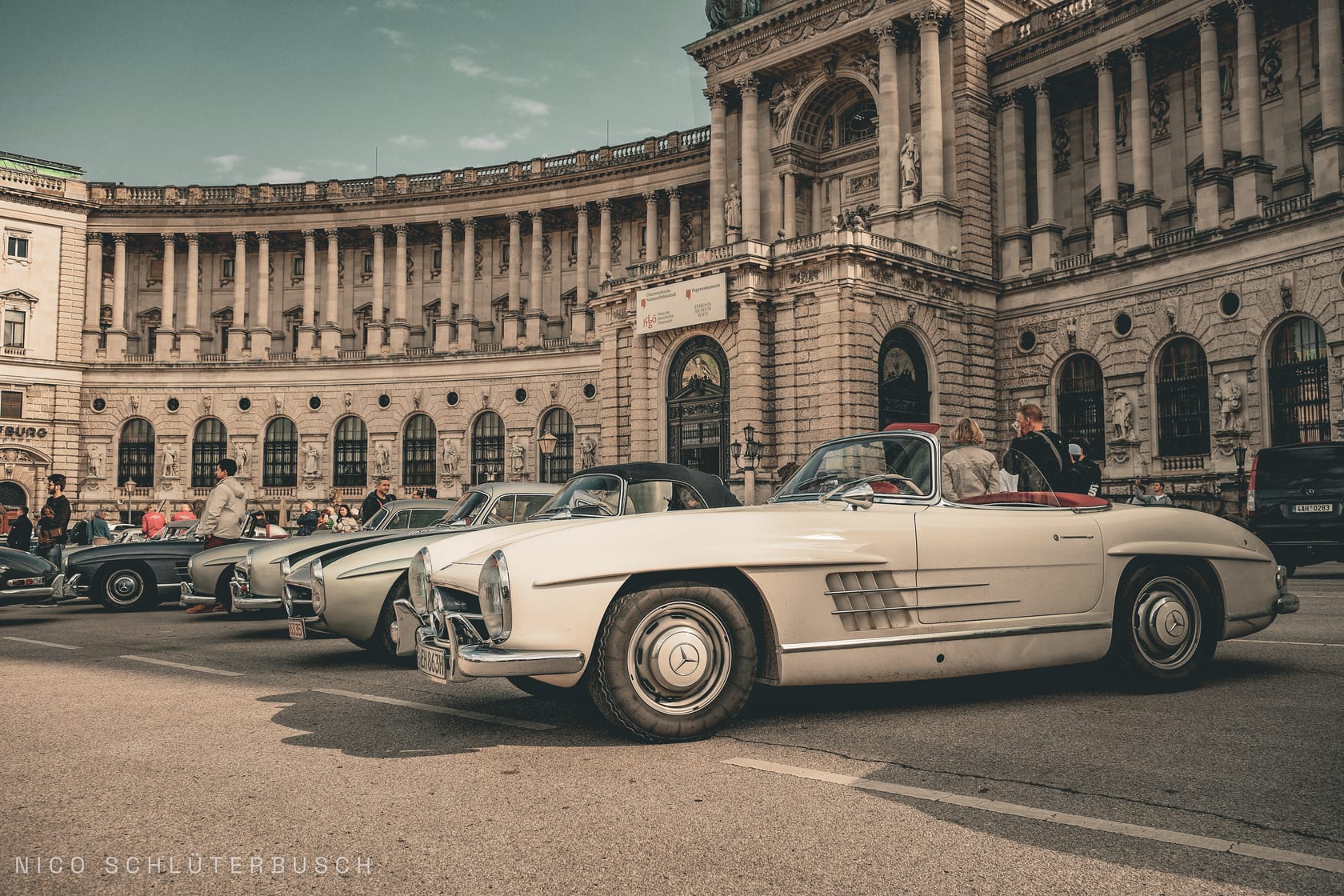 Reihe klassischer Mercedes-Benz 300 SL, vorn ein cremefarbener Roadster mit rotem Interieur, vor der geschwungenen Säulenfassade der Wiener Hofburg